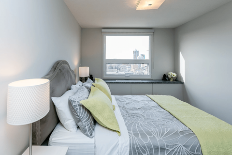 A bedroom with a bed, pillows, a lamp, and a window overlooking a cityscape.