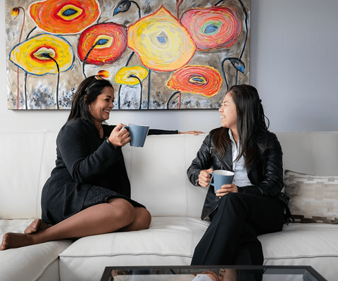 Two women are sitting on a couch, each holding a mug, in front of a colorful floral painting.
