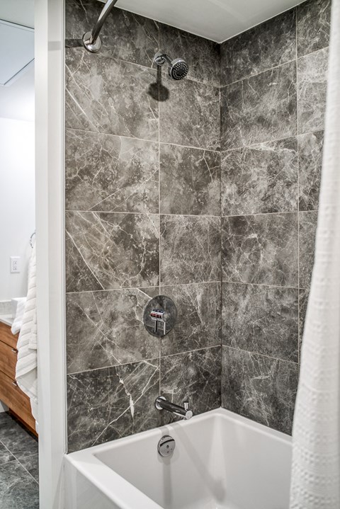 A shower with a marble tile wall and a white tub.
