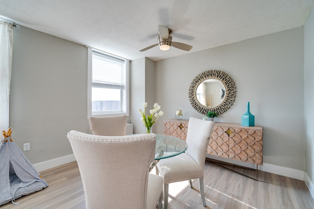 Apartment dining area with a glass table and two chairs, hardwood-style chairs, and windows with natural lighting.