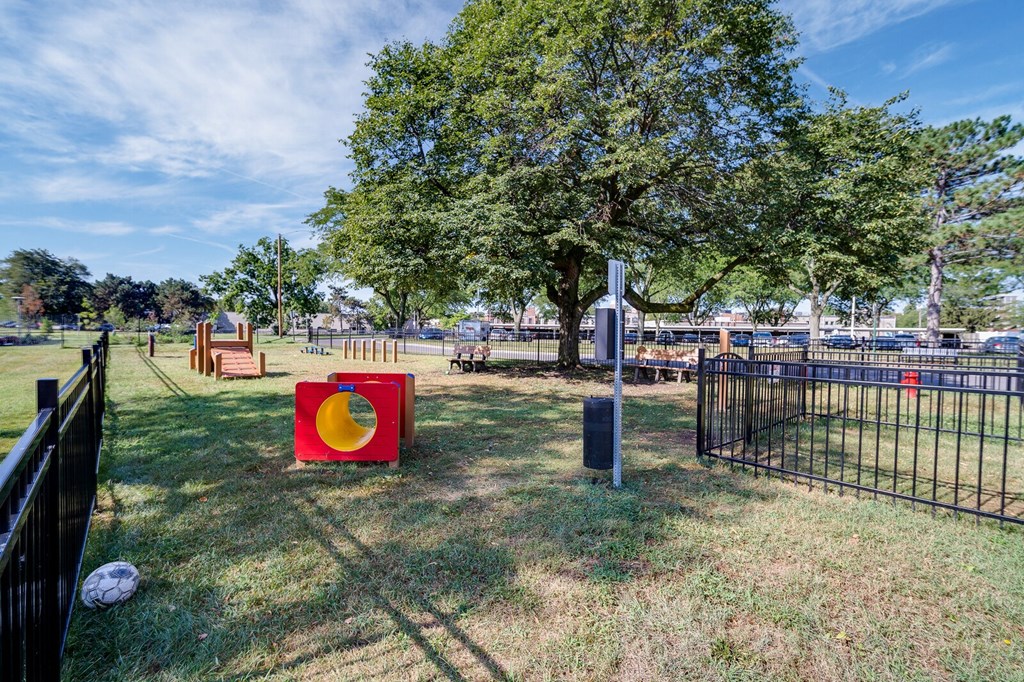 Dayton Towers bark park with a red and yellow play structure, wooden ramp, weaving poles, and a black fence.