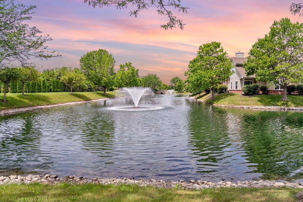 A fountain in the middle of a pond surrounded by trees and a house in the background.