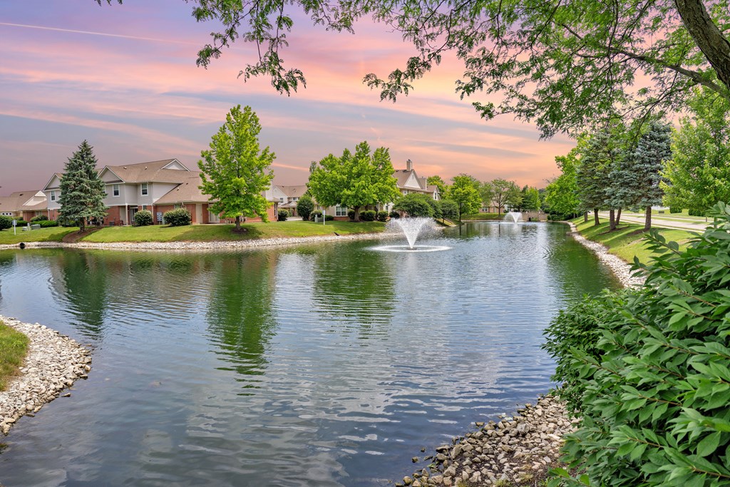 A serene pond with a fountain in the middle surrounded by lush greenery and residential houses in the background.