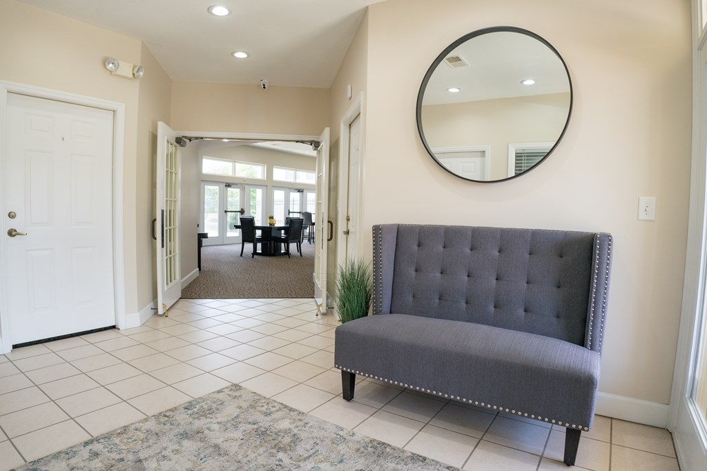 A grey tufted bench sits in a hallway with a round mirror on the wall.