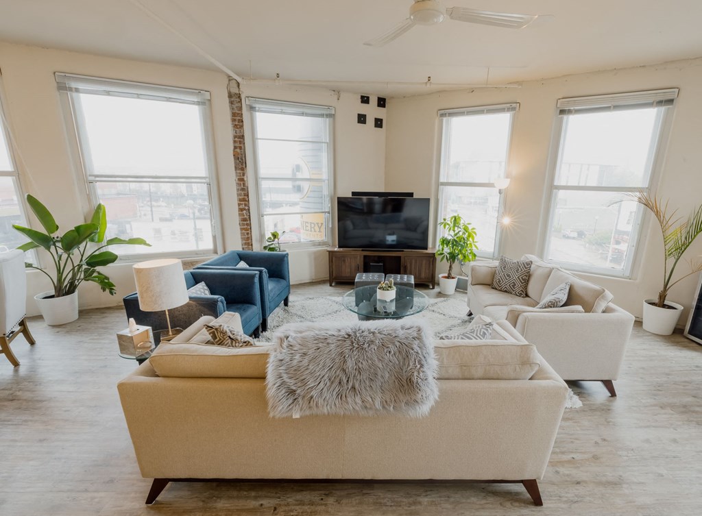 The Barney Power seating area with television, two couches, and two teal chairs. With hardwood-style flooring and four large windows letting in natural lighting.