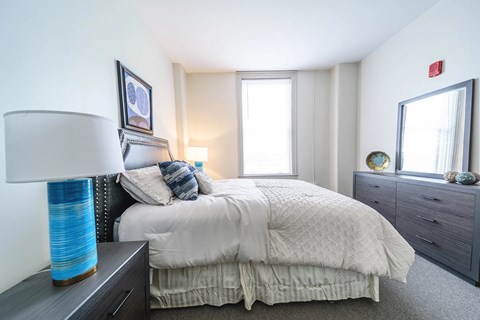 A bedroom with a large bed, a nightstand with a lamp, and a dresser with a mirror.