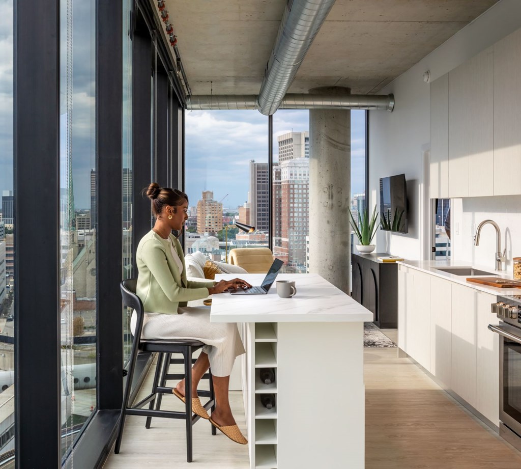 A woman is working on her laptop in a modern kitchen with a view of the city.
