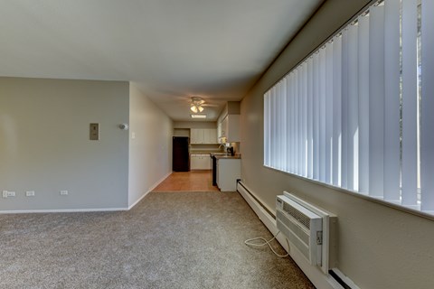 A spacious living room with a carpeted floor, a window with blinds, and a wall-mounted air conditioning unit.