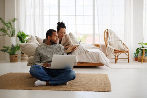 A man and woman are sitting on the floor in a bright room, with the man using a laptop and the woman holding a book.
