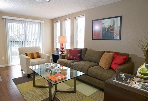 A living room with a glass table and a couch with a red pillow.