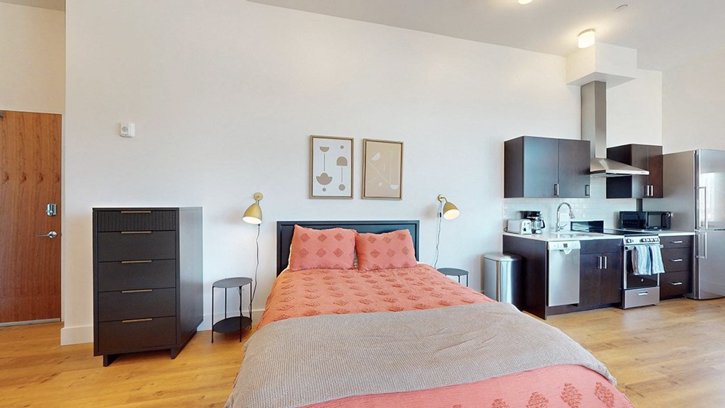 A bedroom with a bed, a dresser, and a kitchen area.