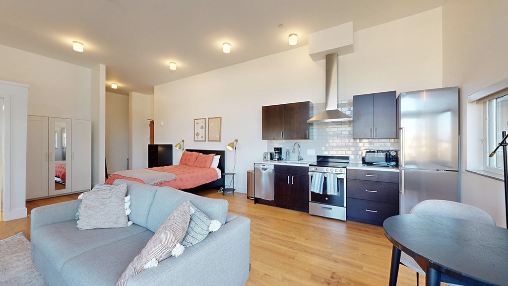 A modern kitchen with a grey couch and a bedroom in the background.