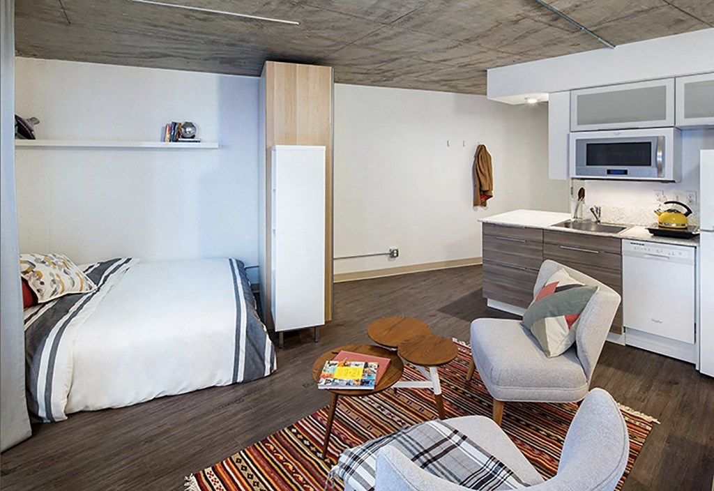 A bedroom with a bed, chair, and a kitchen area.