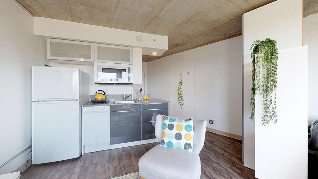 A white chair with a multicolored pillow sits in a kitchen with a white refrigerator and white cabinets.