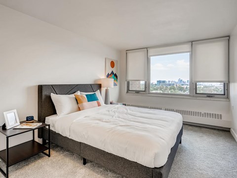 A bedroom with a large bed, a nightstand with a picture frame, and a window with a view of the city.