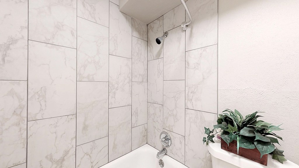 A white bathroom with a marble tile wall and a plant on the tub.