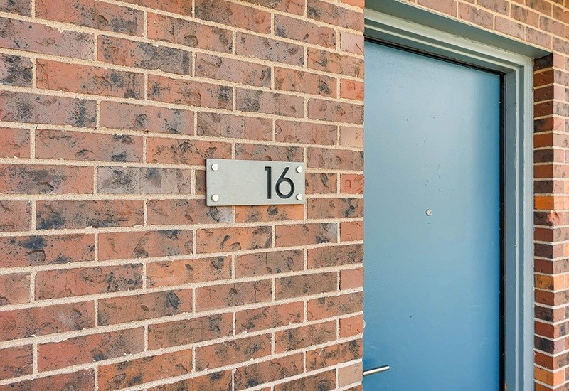 A blue door is on the right side of a brick wall with a white number 16 on a white plaque.