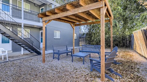 A wooden pergola with a bench and a table is in the foreground of a gravel area.