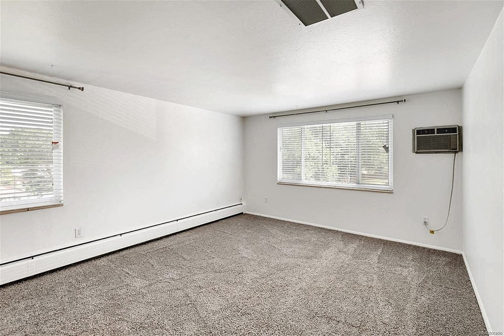 A room with a carpeted floor, a window with blinds, and an air conditioning unit on the wall.