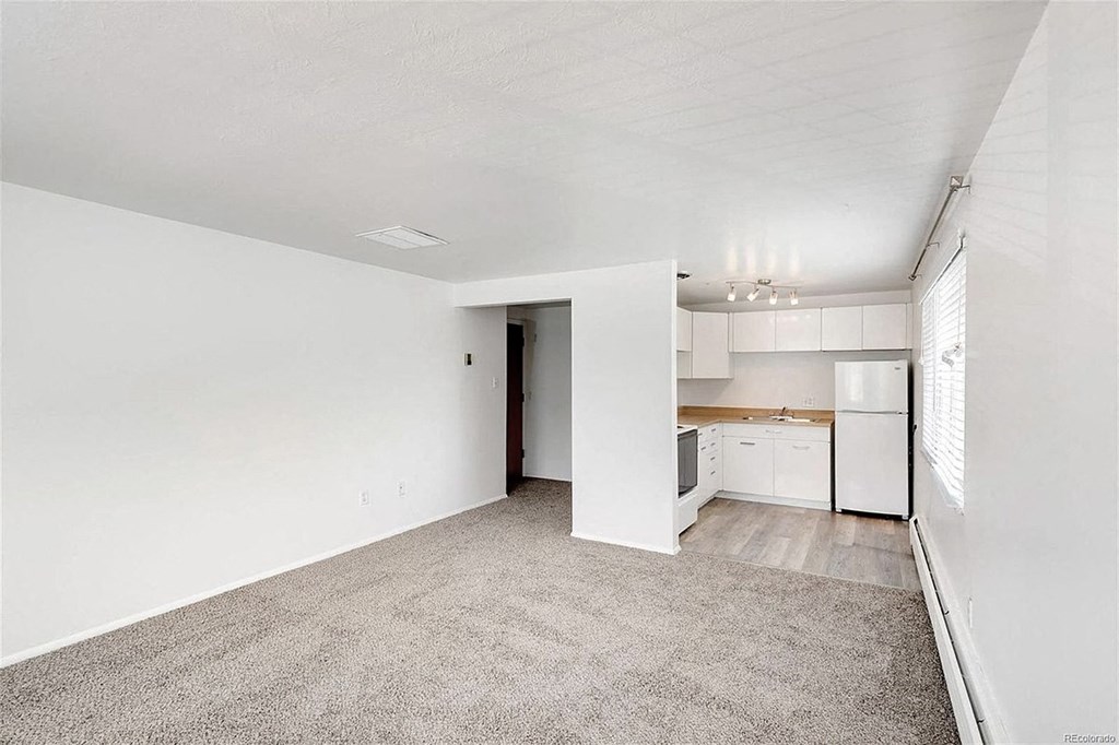 A spacious room with a carpeted floor and a kitchen area visible through an open door.