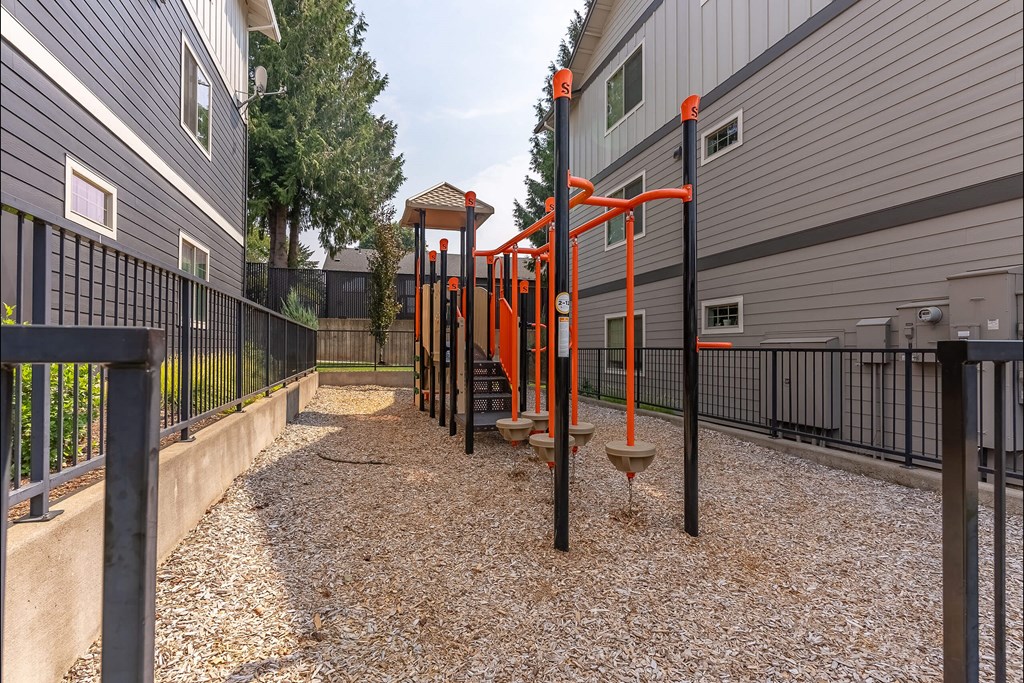 A playground with orange and black equipment is situated between two grey buildings at Sandyplace Apartment Homes, Sandy, OR