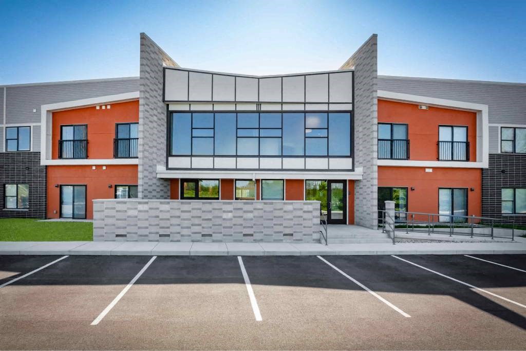 A modern building with orange and grey exterior and a parking lot in front.