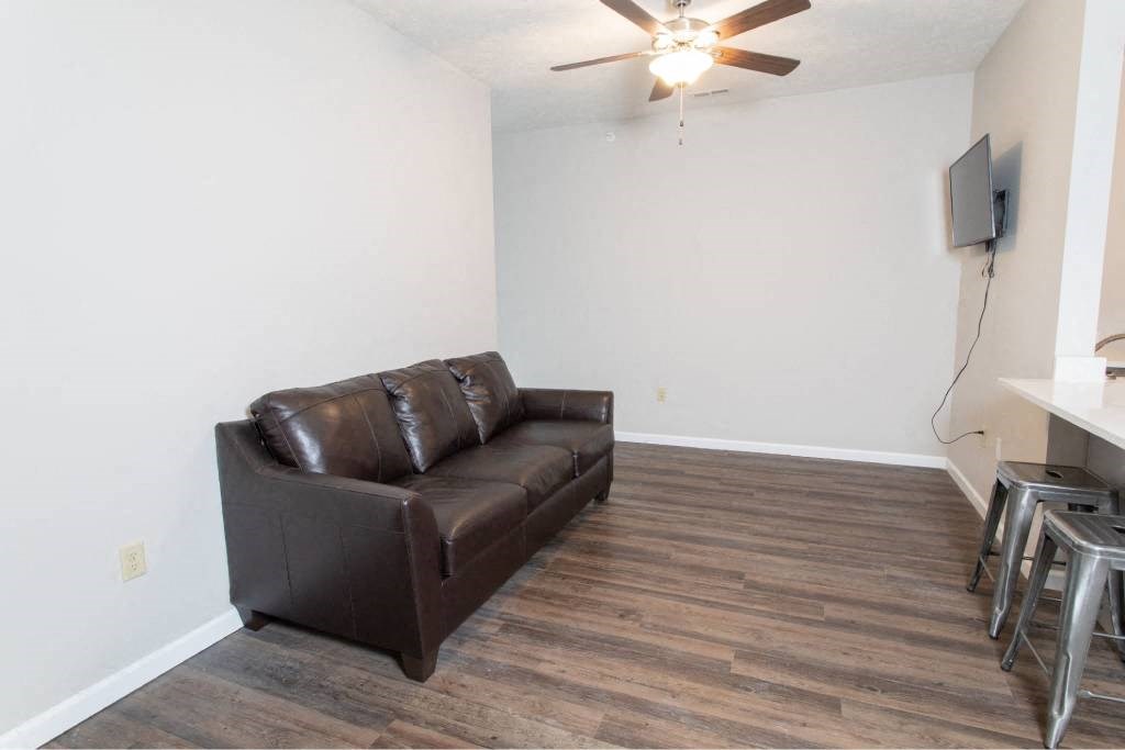 A brown leather couch sits in a room with a ceiling fan and a television mounted on the wall.