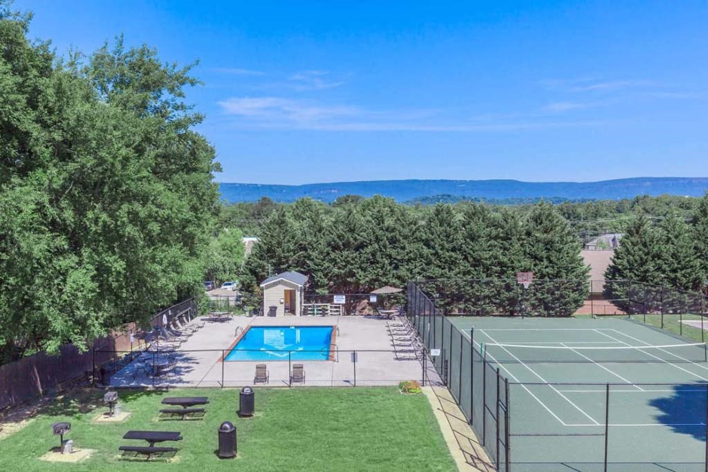 A tennis court is surrounded by a fence and a pool is in the background.