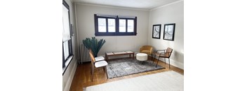 A living room with a brown chair, a white chair, a brown ottoman, a white rug, a brown coffee table, a green plant, and two black and white pictures on the wall.