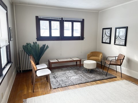A living room with a brown chair, a white chair, a brown ottoman, a white rug, a brown coffee table, a green plant, and two black and white pictures on the wall.