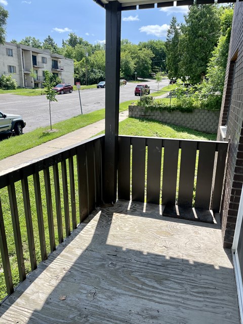 A balcony with a wooden floor and a black railing overlooks a street with cars and buildings.