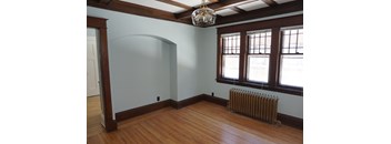 A room with a wood floor, a white wall, a window with a brown frame, and a chandelier hanging from the ceiling.