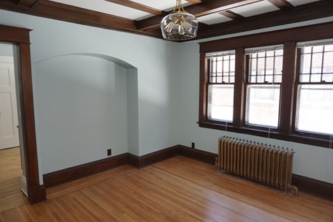 A room with a wood floor, a white wall, a window with a brown frame, and a chandelier hanging from the ceiling.