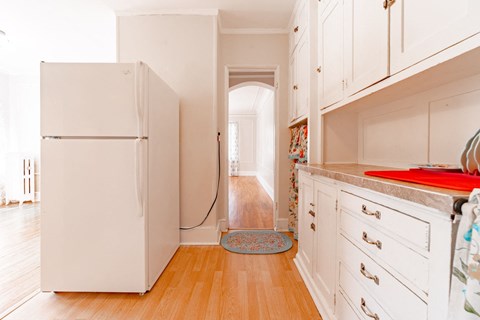 A white refrigerator in a kitchen with wooden floors and white cabinets.