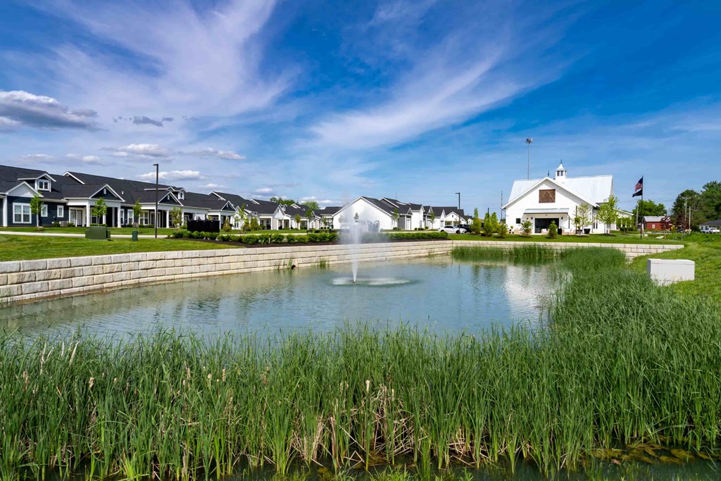 A white building with a flag on top stands behind a pond with a fountain in the middle.