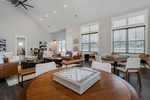 A large wooden table with a checkerboard in the middle is in the foreground of a spacious room with multiple seating areas and large windows.