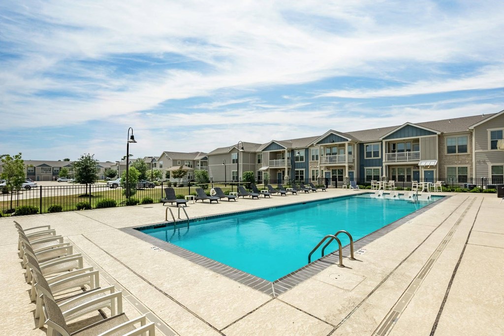A large swimming pool surrounded by lounge chairs in front of apartment buildings.