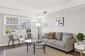 A living room with a grey couch, a glass table, and a window with a view of the street at The Preserve at Pine Valley Apartments, Wilmington, 28412