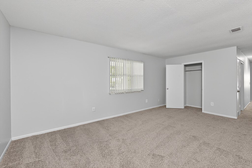 A large empty room with carpeted floor and a window with blinds at Brackenbrook Apartments, North Charleston 29418