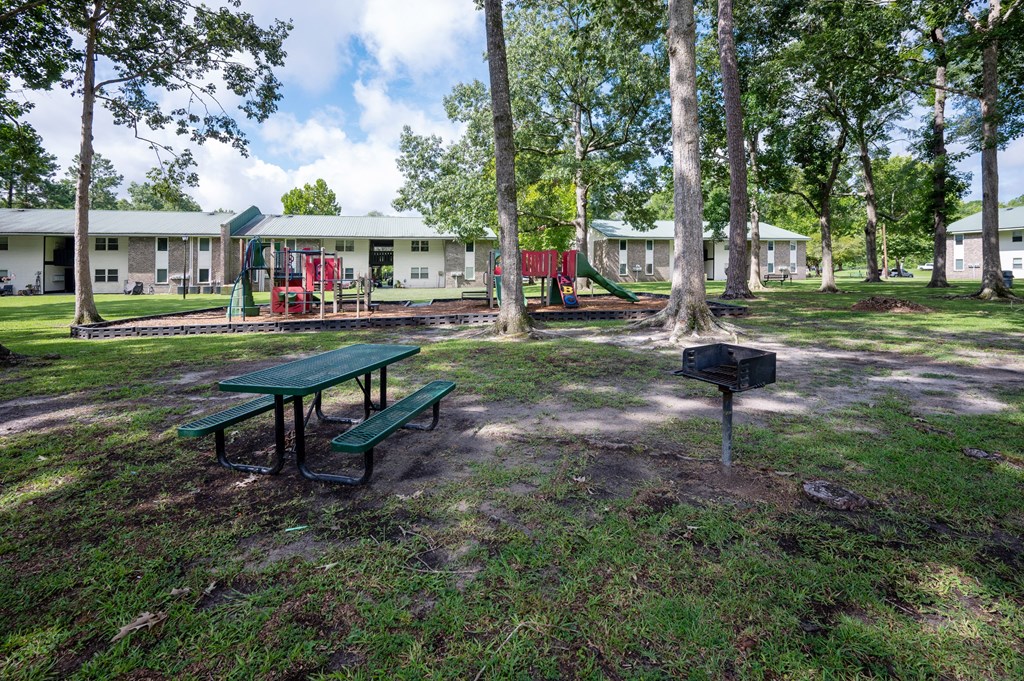 A green picnic table sits in a grassy area in front of a playground and a building at Sawbranch Apartments, South Carolina, 29485