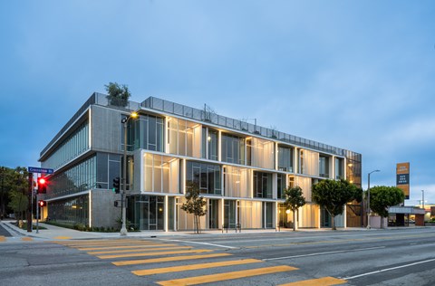 A modern building with a glass facade is illuminated from within and surrounded by a yellow crosswalk.