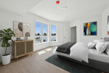 A bedroom with a bed, a plant, and a window overlooking a tree-filled street.