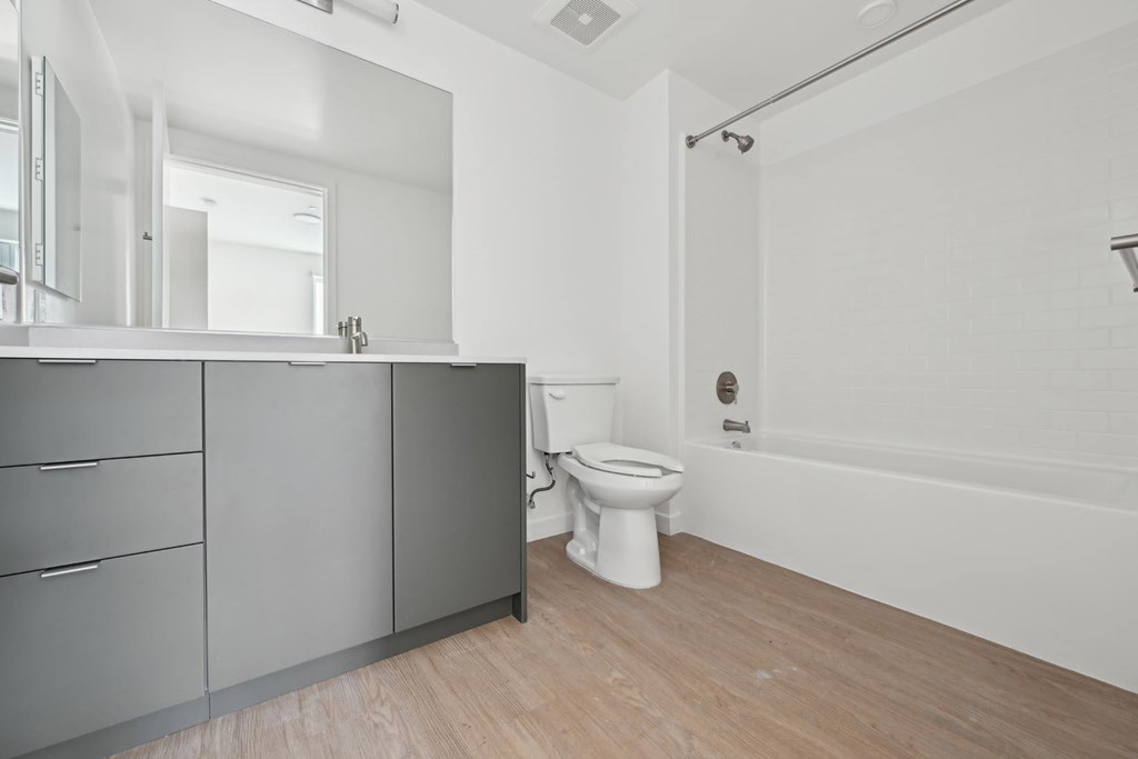 A white toilet sits in a bathroom with a grey cabinet and a white bathtub.