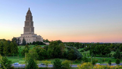 A large, towering building with a spire is surrounded by trees and greenery.