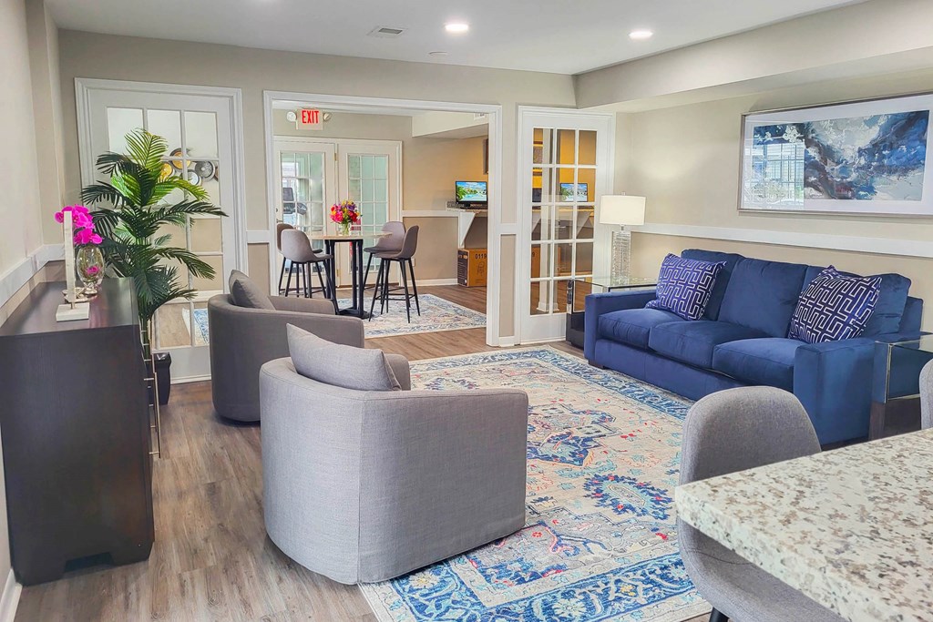A living room with a grey sofa, a grey armchair, a grey ottoman, a wooden side table, a glass table with chairs, a rug, and a painting on the wall.