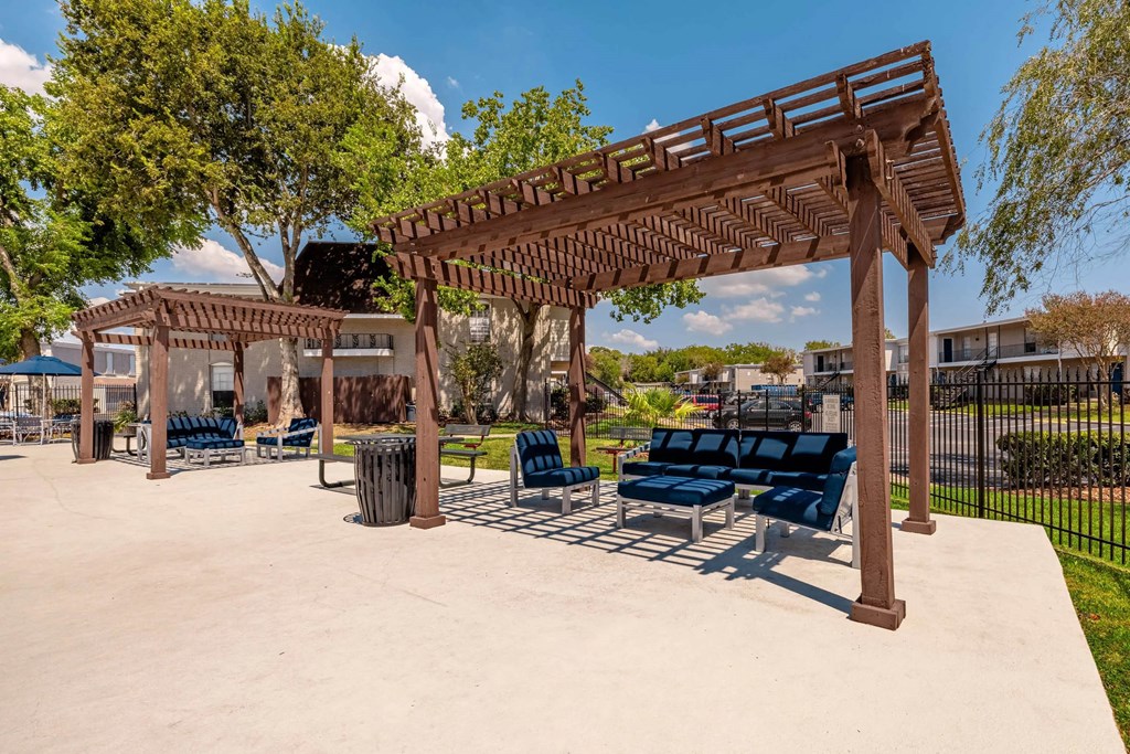 A wooden pergola with blue cushioned seating is in the foreground of a sunny park.
