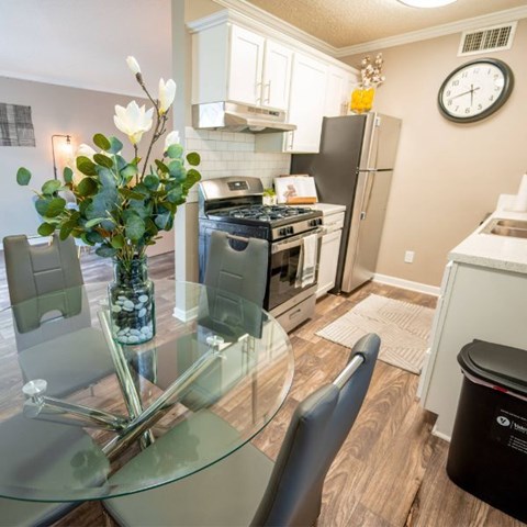 A kitchen with a glass table and chairs, a vase with flowers, and a clock on the wall.