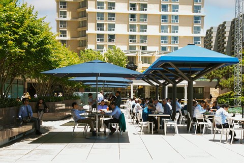 People are sitting at tables under blue umbrellas in a courtyard.