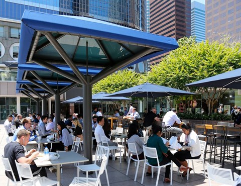 People are sitting at tables under blue umbrellas in an outdoor dining area.