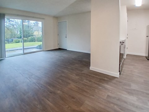 A spacious room with wooden flooring and a view of a green outdoor area through the windows.